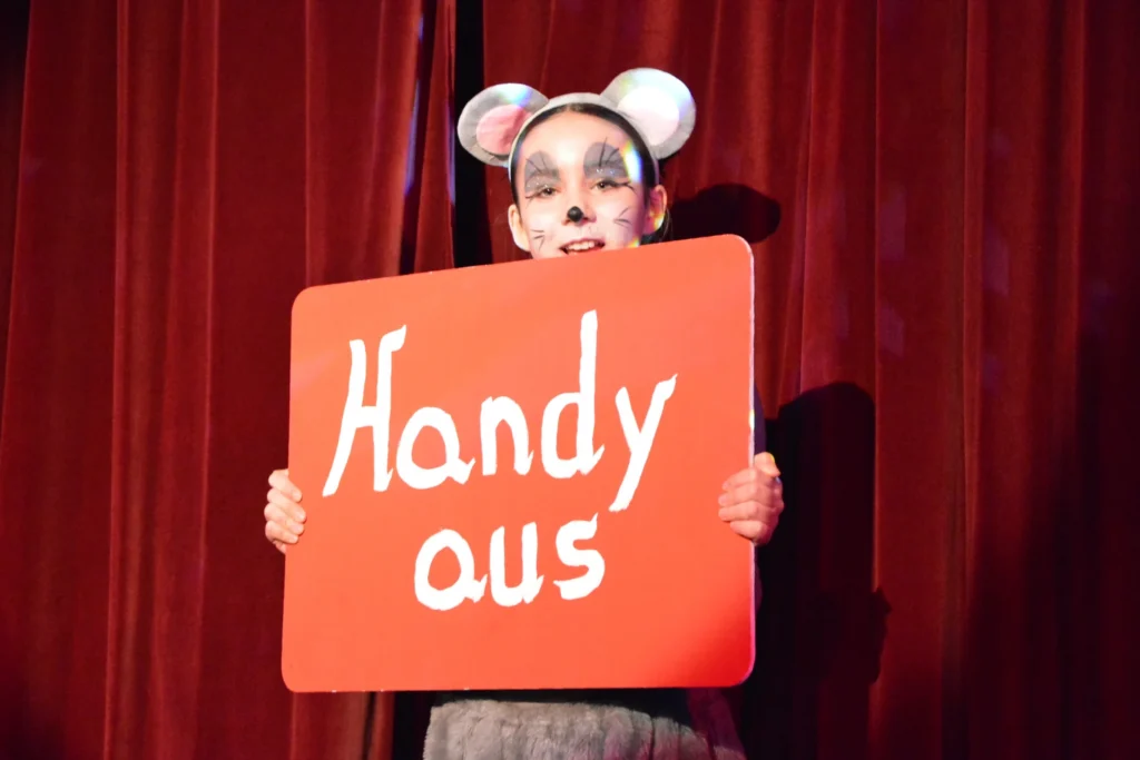 Eine Figur im Mäusekostüm hält auf der Bühne ein rotes Schild mit der Aufschrift Handy aus