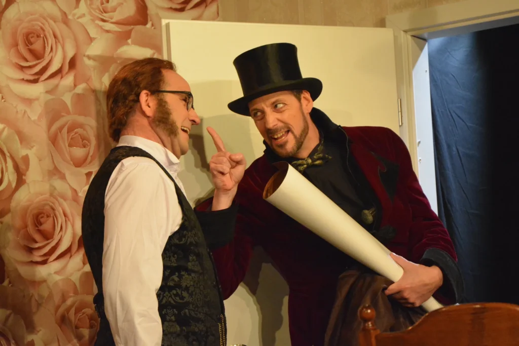 Ein Mann mit Zylinder, gerolltem Plan und erhobenem Zeigefinger spricht zu einem Mann in Weste