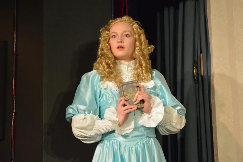 Eine blonde Darstellerin im hellblauen Kleid hält überrascht ein kleines Buch an die Brust