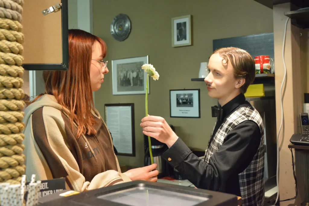 Ein Darsteller in karierter Weste bietet einer Frau am Tresen im Theaterfoyer eine weiße Blume an