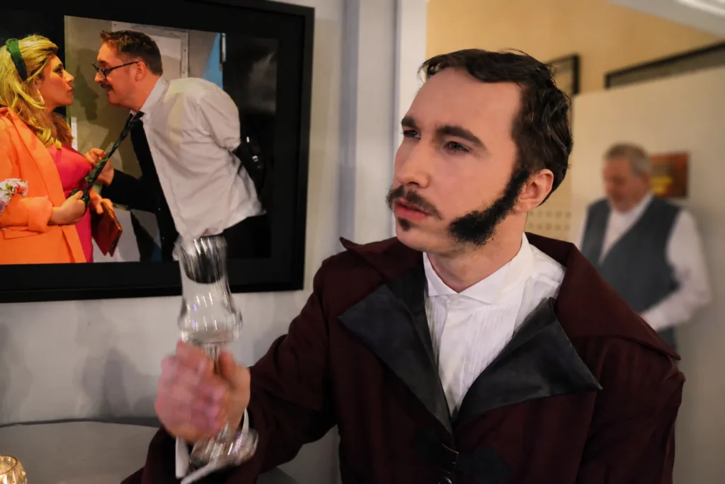 Ein junger Mann im burgunderfarbenen Mantel hebt im Theaterfoyer ein Glas